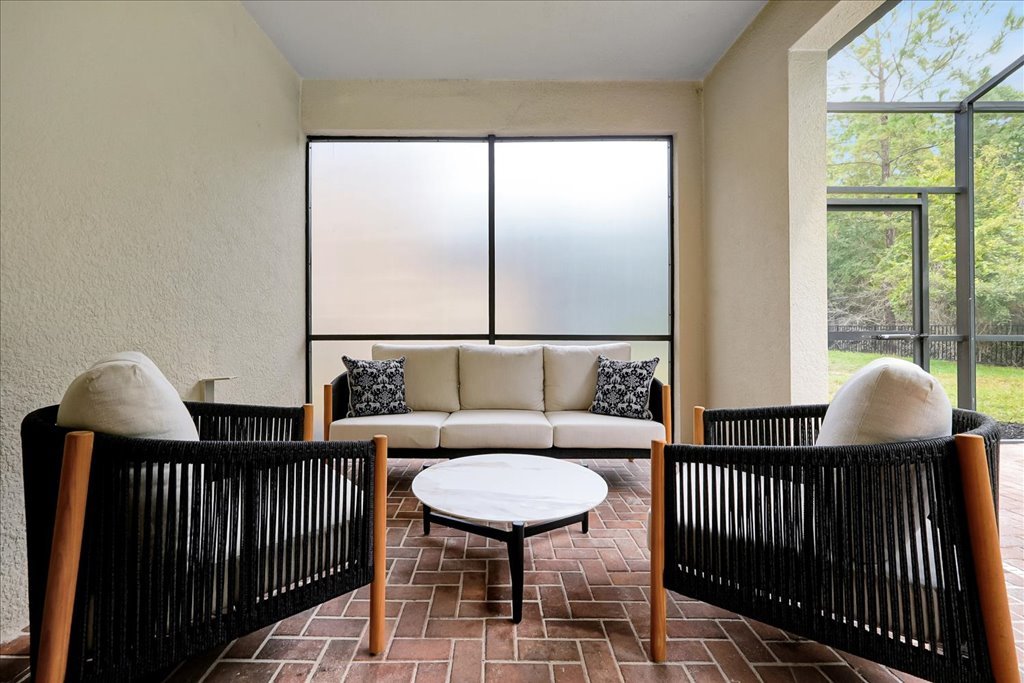 A cozy sunroom with a white sofa, two black and white armchairs, a round white coffee table, and large windows showing greenery outside.