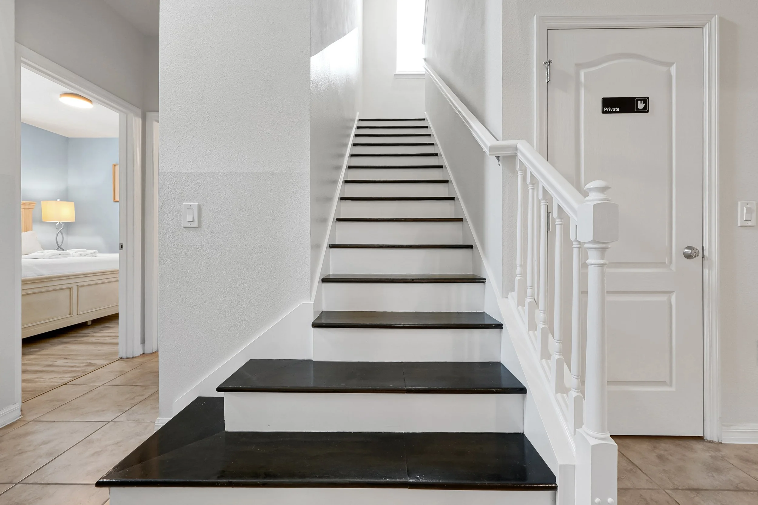 Interior view of a staircase with black steps and white risers, white handrail, and white walls. To the right, there is a door with a "Private" sign and a hand icon. To the left, an open doorway leads to a bedroom with a bed, nightstand, and lamp.