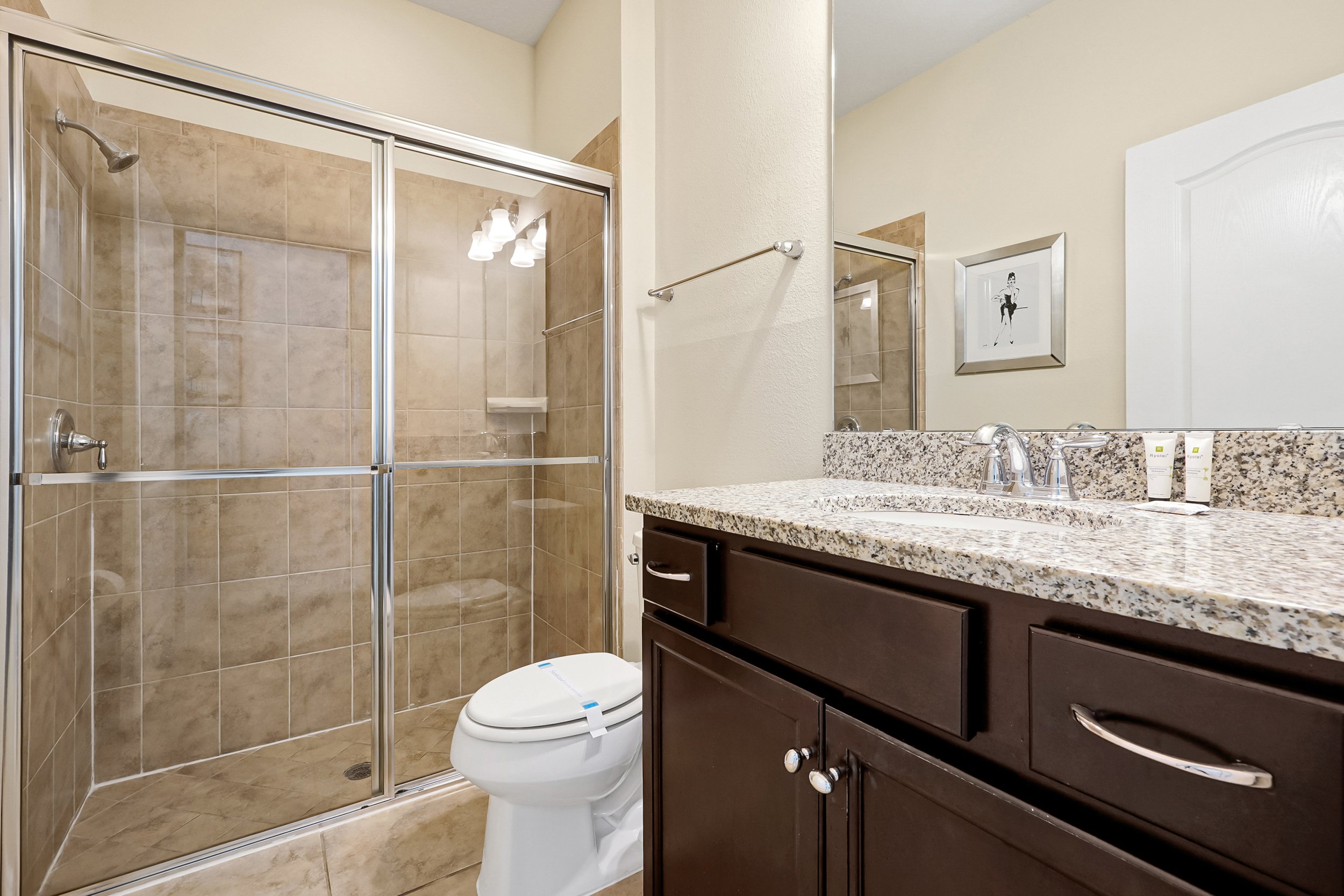 Bathroom with a glass-enclosed shower, toilet, and a dark wood vanity with a granite countertop. There is a framed picture on the wall.