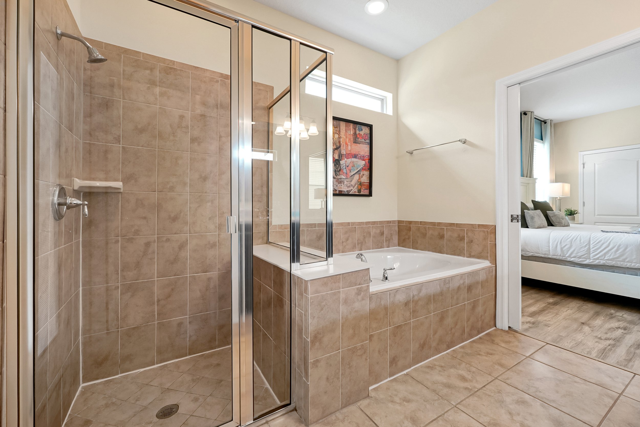 A bathroom with a glass shower enclosure, a bathtub, beige tile flooring and walls, a framed abstract artwork, and an opening leading to a bedroom with a bed, pillows, a nightstand, and a lamp.