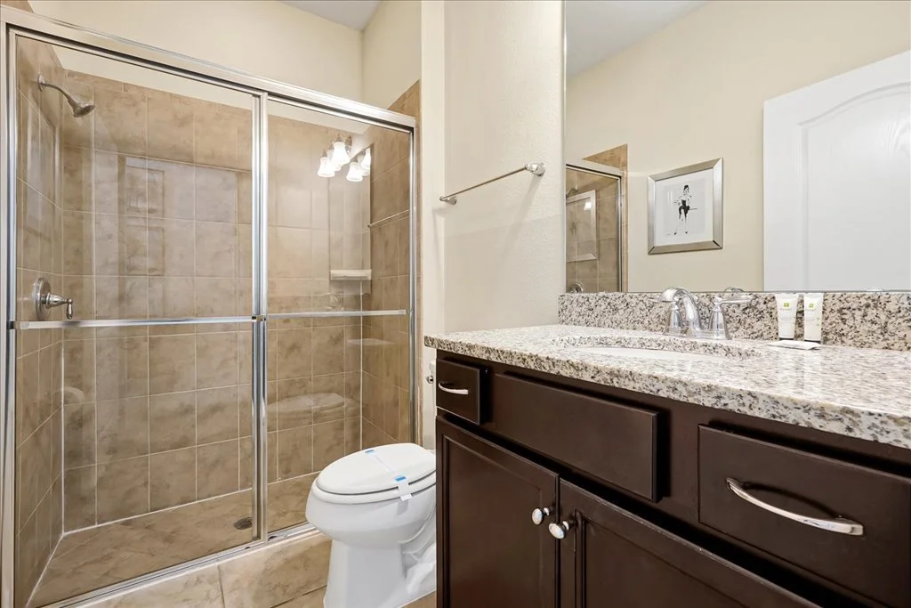 Bathroom with a glass-enclosed shower, toilet, granite countertop sink, mirror, and framed picture on the wall.