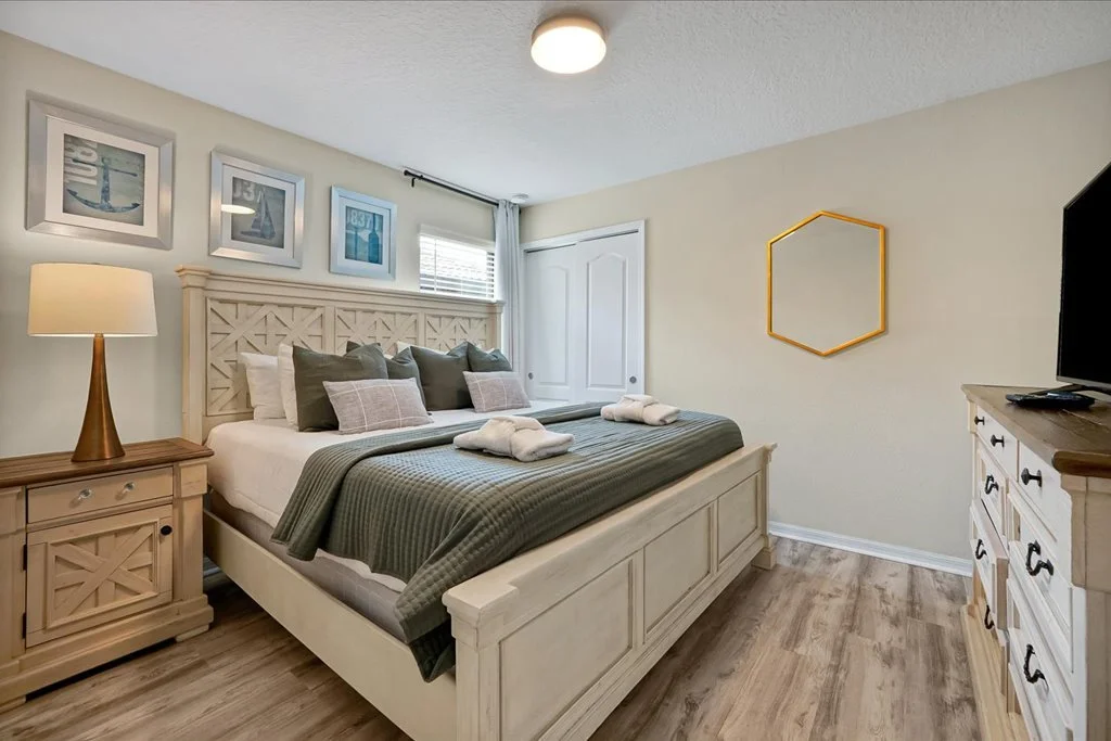 A bedroom with a white wooden bed, green bedding, pillows, a nightstand with a lamp, framed pictures of boats above the bed, a dresser with a TV, and a yellow hexagonal mirror on the wall.