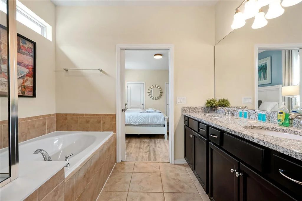 Bathroom with a bathtub and a double sink vanity, connected to a bedroom with a bed and nightstand.