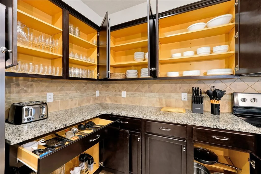 Kitchen cabinet open with dishes and glassware, black cabinets, granite countertop, toaster, knife set, utensils, stovetop, and a partially opened drawer with kitchen utensils visible.