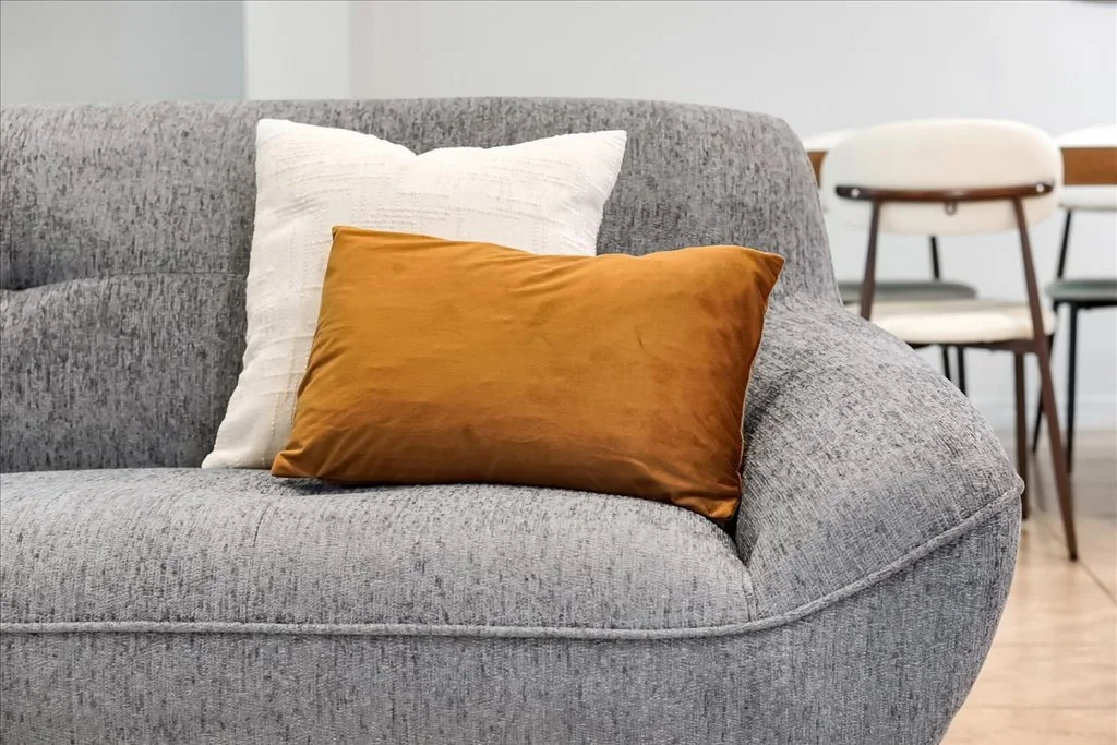A gray upholstered sofa with a white cushion and a brown cushion, with two chairs visible in the background.