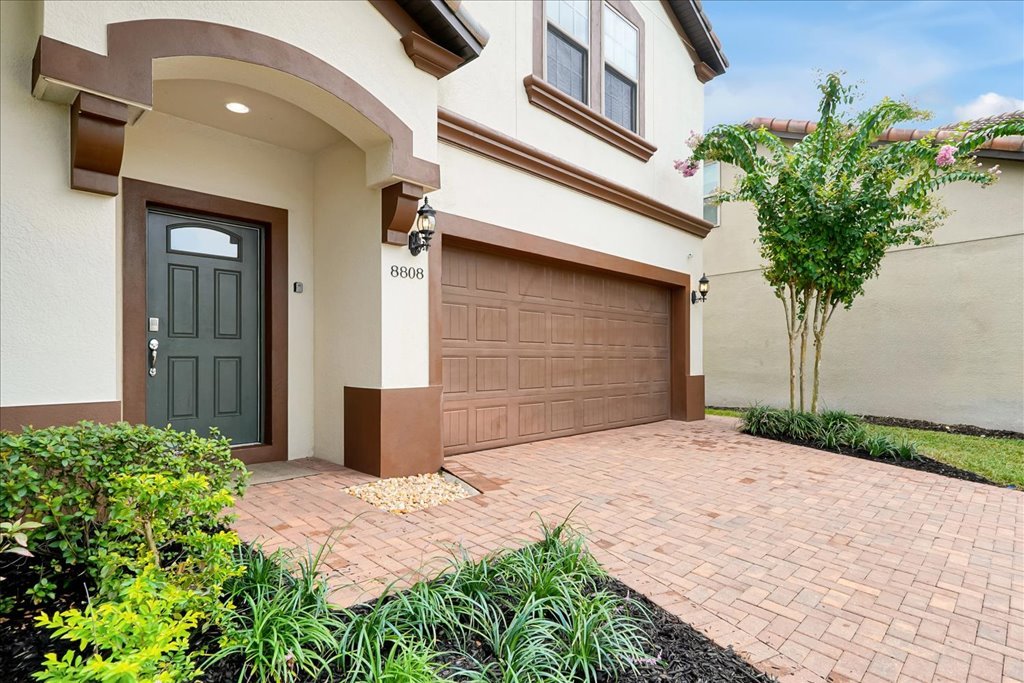 Street view of a modern house with a dark green front door, brown garage door, and landscaped yard with green shrubs, small trees, and brick pavement.