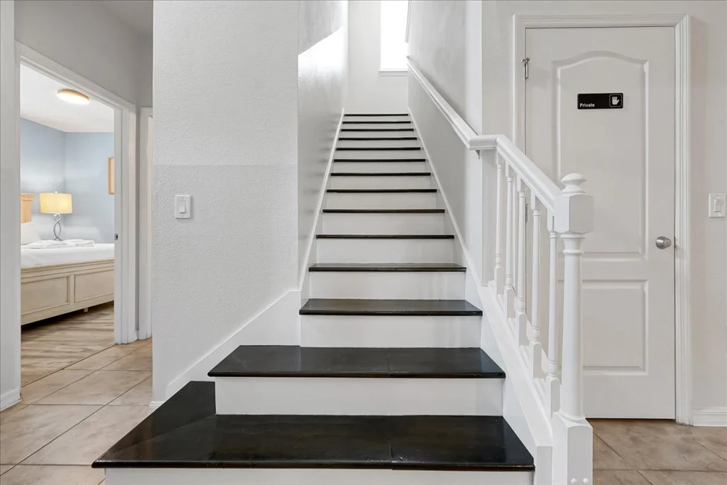 White staircase with dark-colored steps and white railing, leading to the upper floor in a house. To the right is a door with a 'Private' sign, and to the left is an entrance to a bedroom with a bed and bedside lamp.