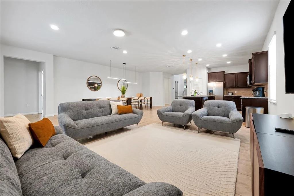 Open-concept living room and kitchen with gray sofas, armchairs, a white rug, dark wood cabinets, and modern lighting.