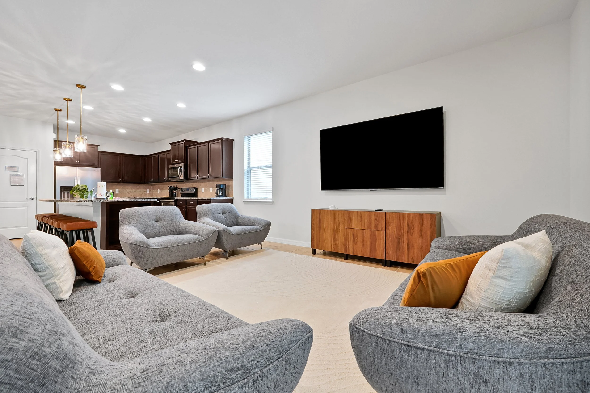 Living room with gray fabric sofas, decorative pillows, wooden TV stand, large flat-screen TV, and kitchen area with pendant lights and cabinetry.