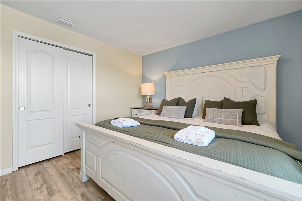 A neatly made bed with multiple pillows and folded towels, a bedside table with a lamp, a closed closet door, and hardwood flooring in a bedroom with light-colored walls and a decorative headboard.