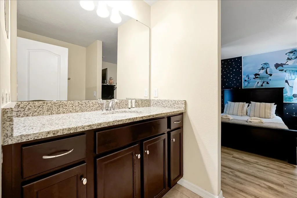 Bathroom vanity with granite countertop, dark wood cabinets, and a large mirror. Part of a bedroom with Star Wars-themed wall decor and bed with striped pillows is visible in the background.