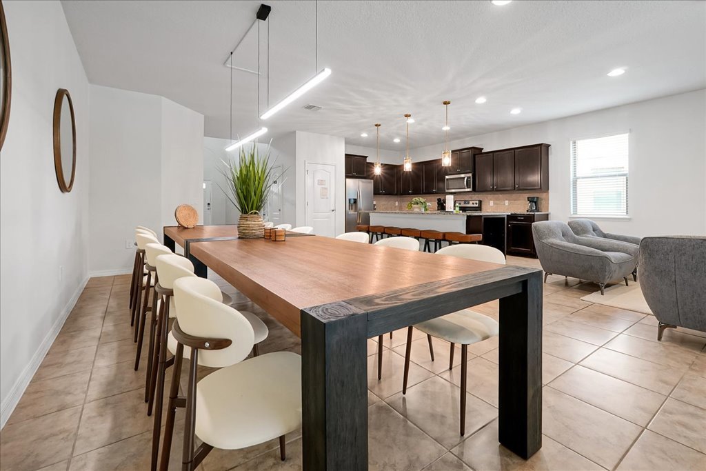 Open-plan kitchen and dining area with a large wooden table, white chairs, gray armchairs, dark kitchen cabinets, and pendant lights.