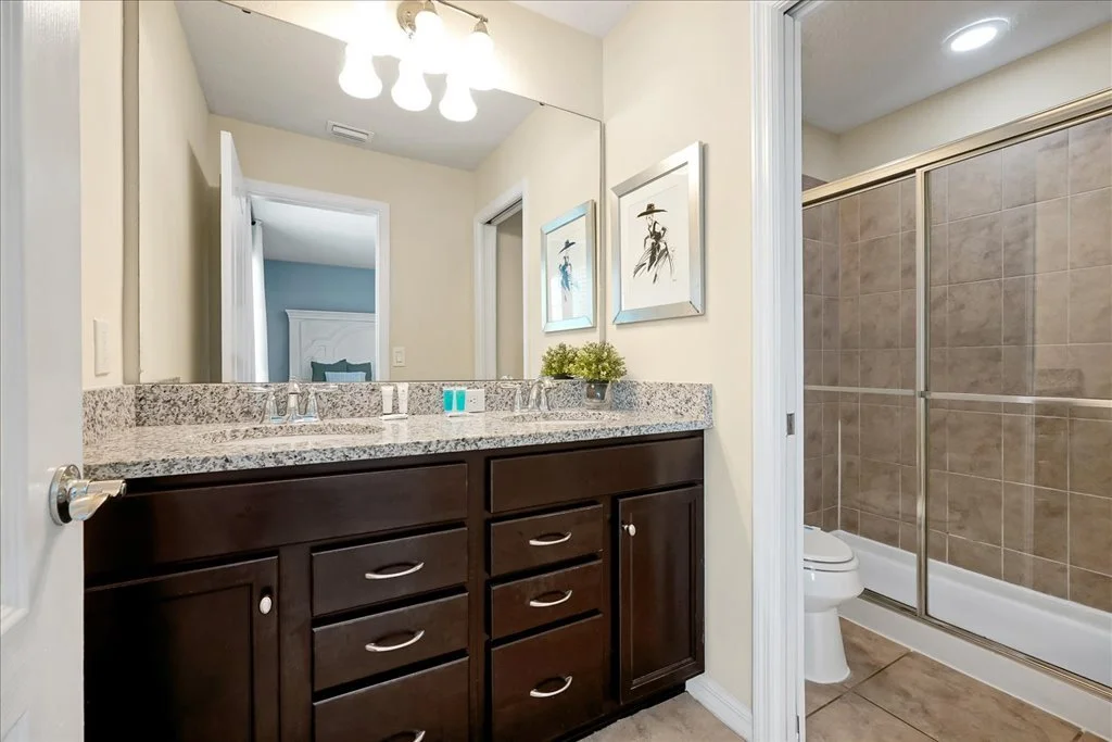 Bathroom with a double vanity, granite countertop, dark wooden cabinets, and a large mirror. Two framed pictures hang on the wall. To the right is a shower with sliding glass doors and brown tiles. A toilet is adjacent to the shower.