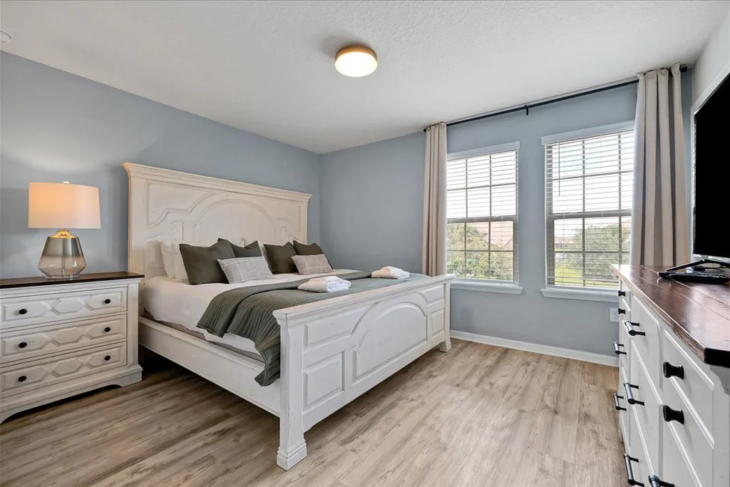 A bedroom with a white bed, dark pillows, light blue walls, and two windows with white blinds and curtains.