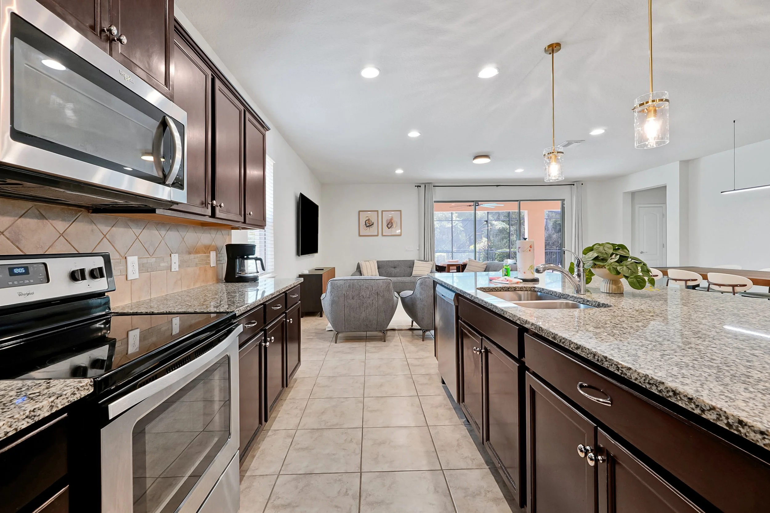 A modern open-concept kitchen with dark wood cabinets, stainless steel microwave, electric stove, granite countertops, tiled floor, and a view into the living room with a window, sofa, wall-mounted TV, and dining area with chairs and large window.