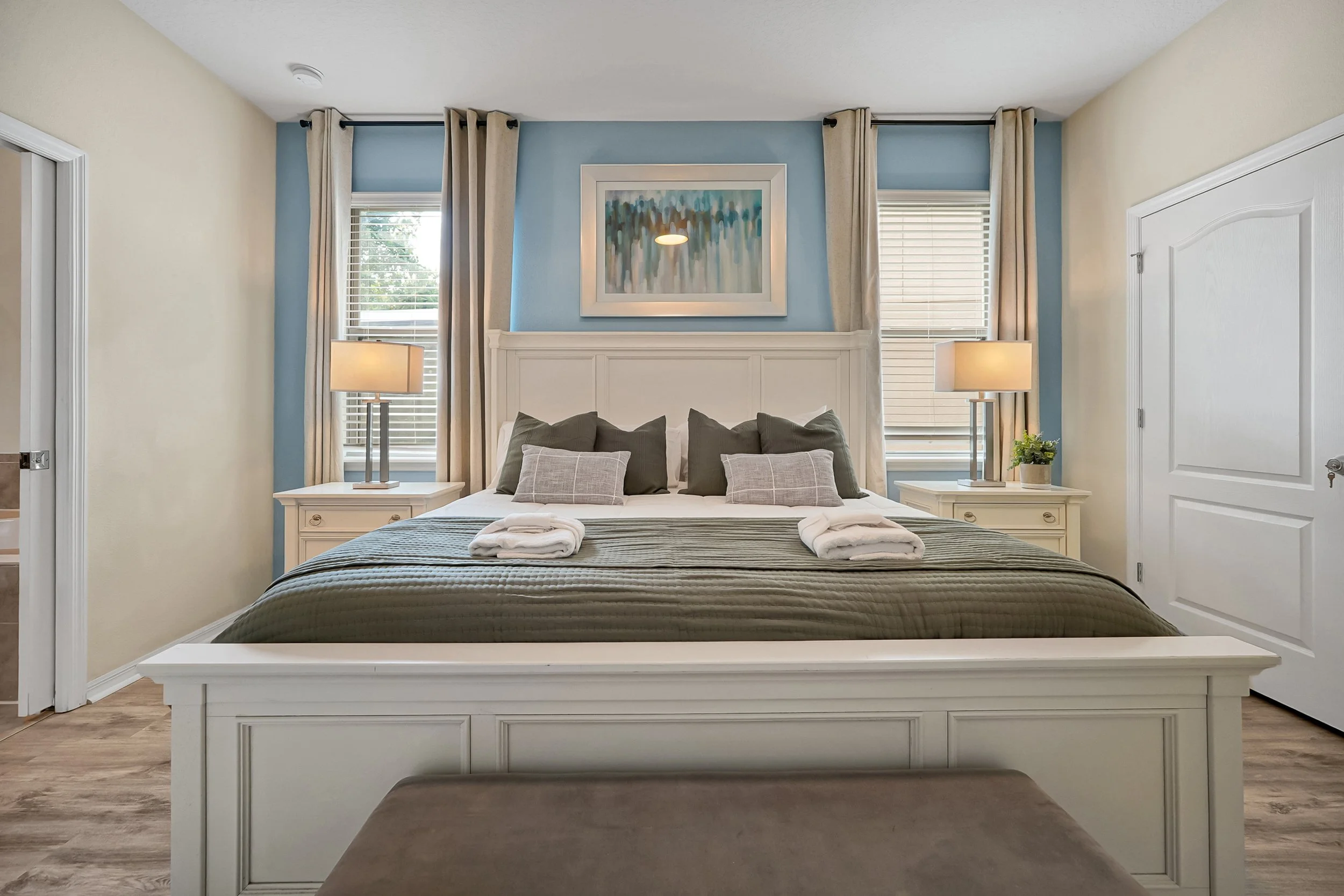 A neatly made bed with dark green and light gray pillows, with two folded towels on top, in a bedroom with light blue and cream walls, two windows with white blinds and curtains, and nightstands with lamps on each side of the bed.