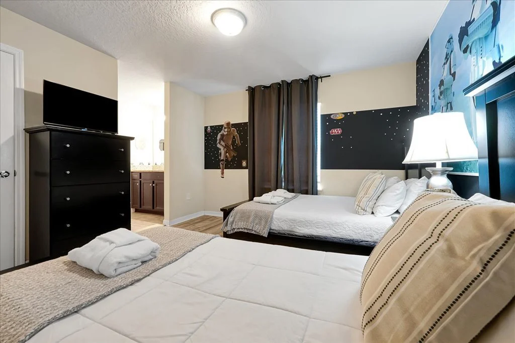 A hotel room with two beds, Star Wars-themed wall decor, a nightstand with a lamp, a dresser with a flat-screen TV, and a bathroom visible through an open door.