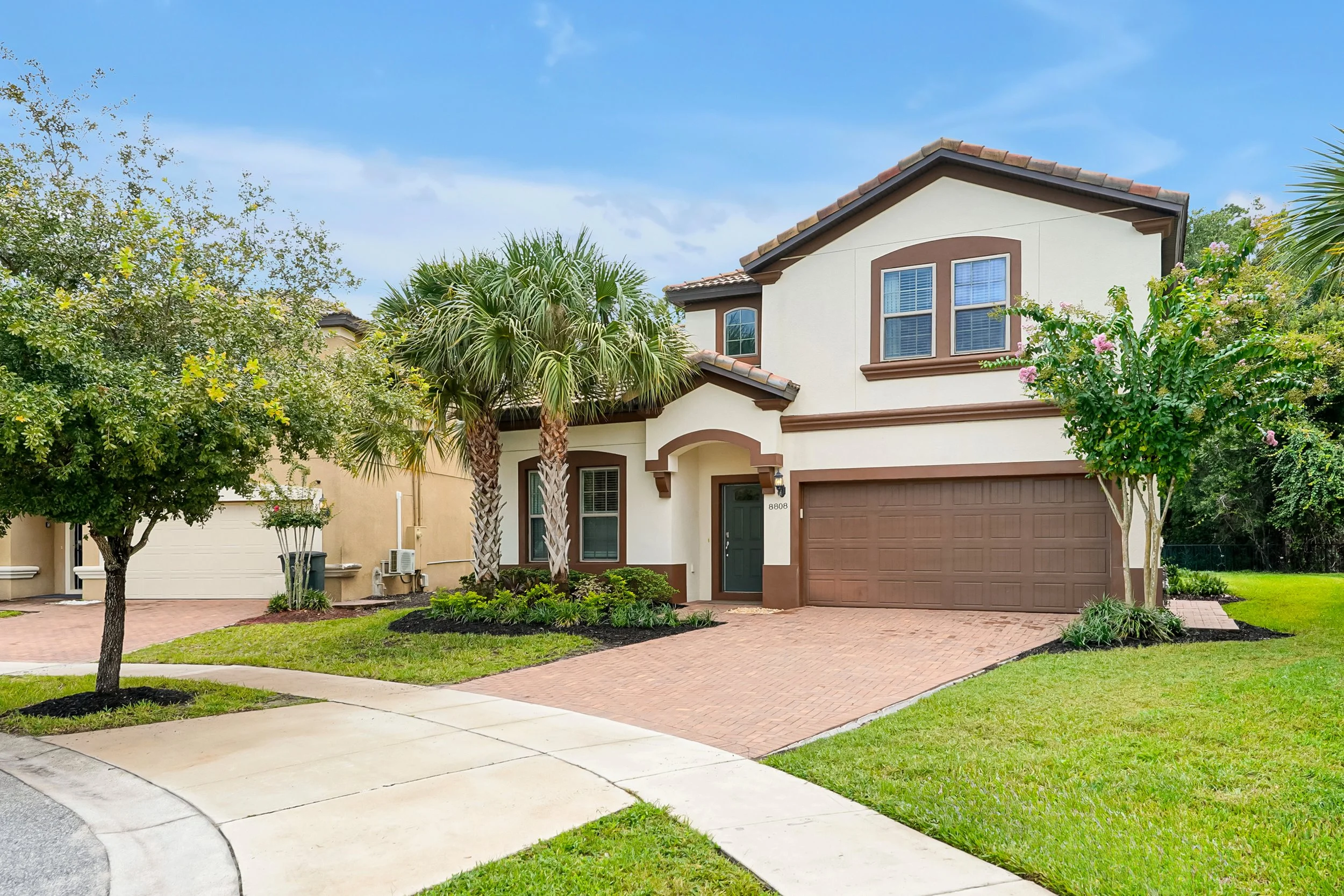 A two-story suburban house with a brown garage door, light cream exterior, and brown trim. The house has roof tiles and several windows with white blinds. The front yard features a palm tree, a small blooming tree, and well-maintained grass and plant