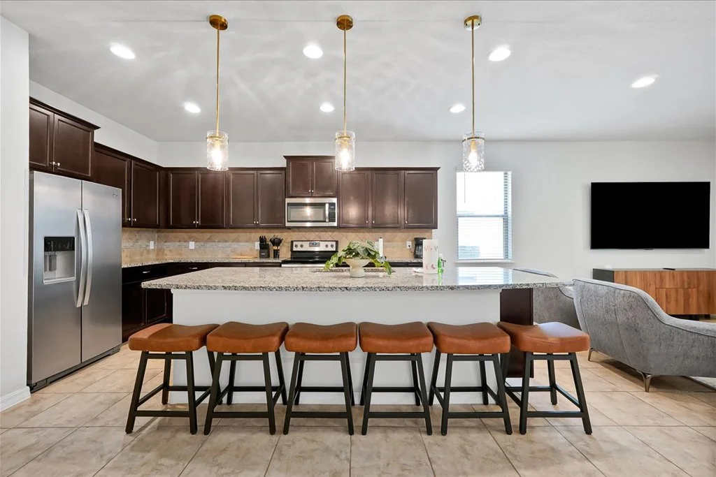 Modern kitchen with dark wood cabinets, stainless steel refrigerator, and a granite island with six brown leather stools. Pendant lights hanging above the island, white walls, a sliding window, and a wall-mounted TV.