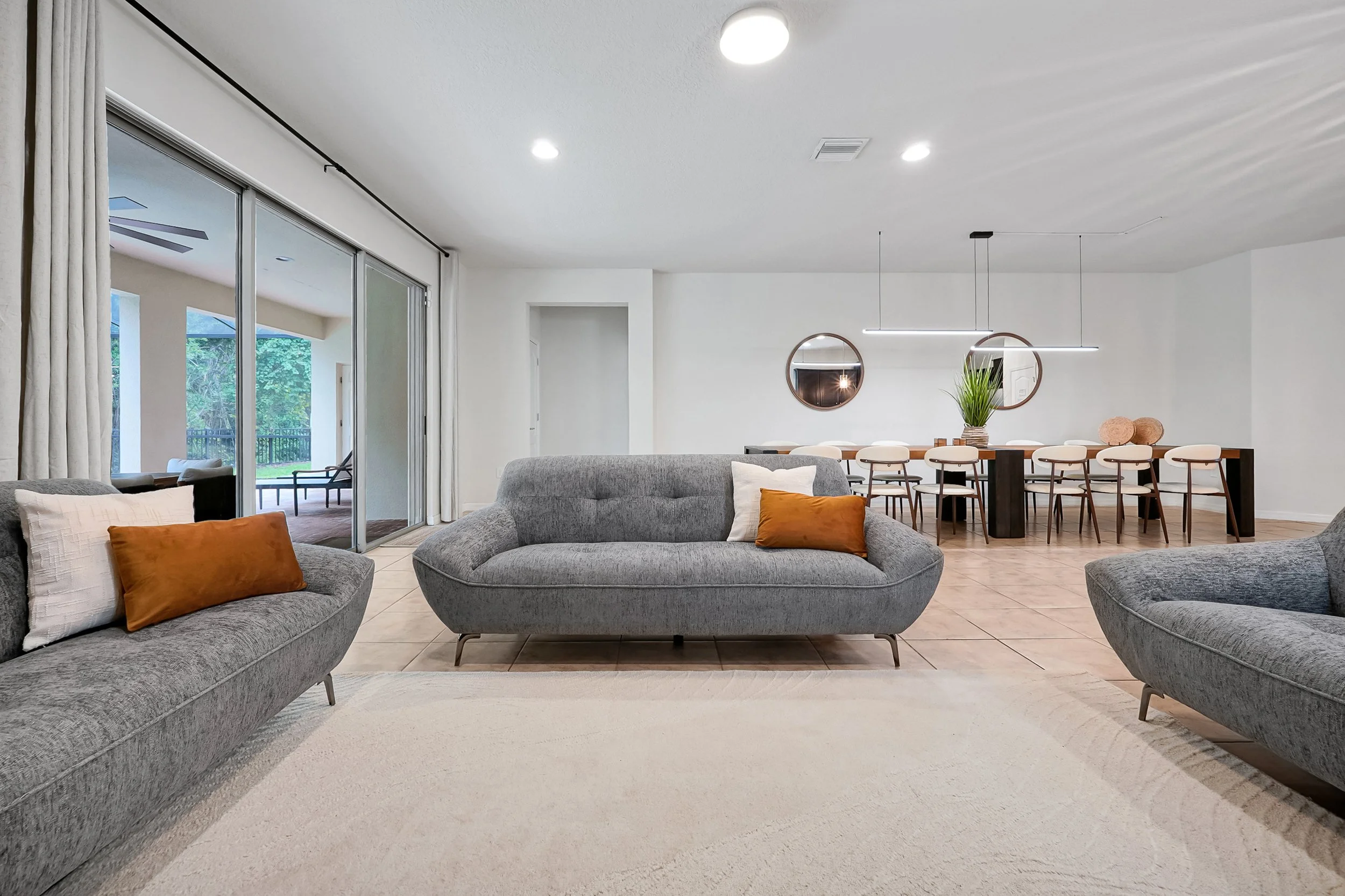 Modern living room with three gray sofas, assorted pillows, a dining area with a long table and chairs, large sliding glass doors leading to an outdoor patio, and two circular wall mirrors.