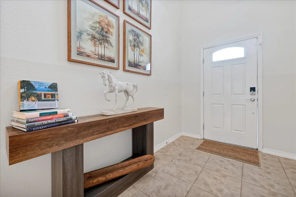 Entryway with a white door and a window, a beige tiled floor, a wooden console table with a stack of books and a decorative horse figurine, and framed landscape paintings on the wall.
