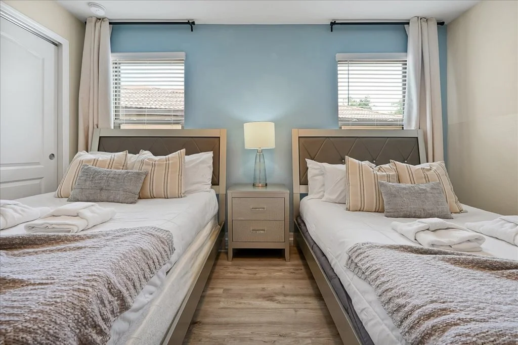 A bedroom with two beds, a nightstand, and two windows with blinds and curtains.