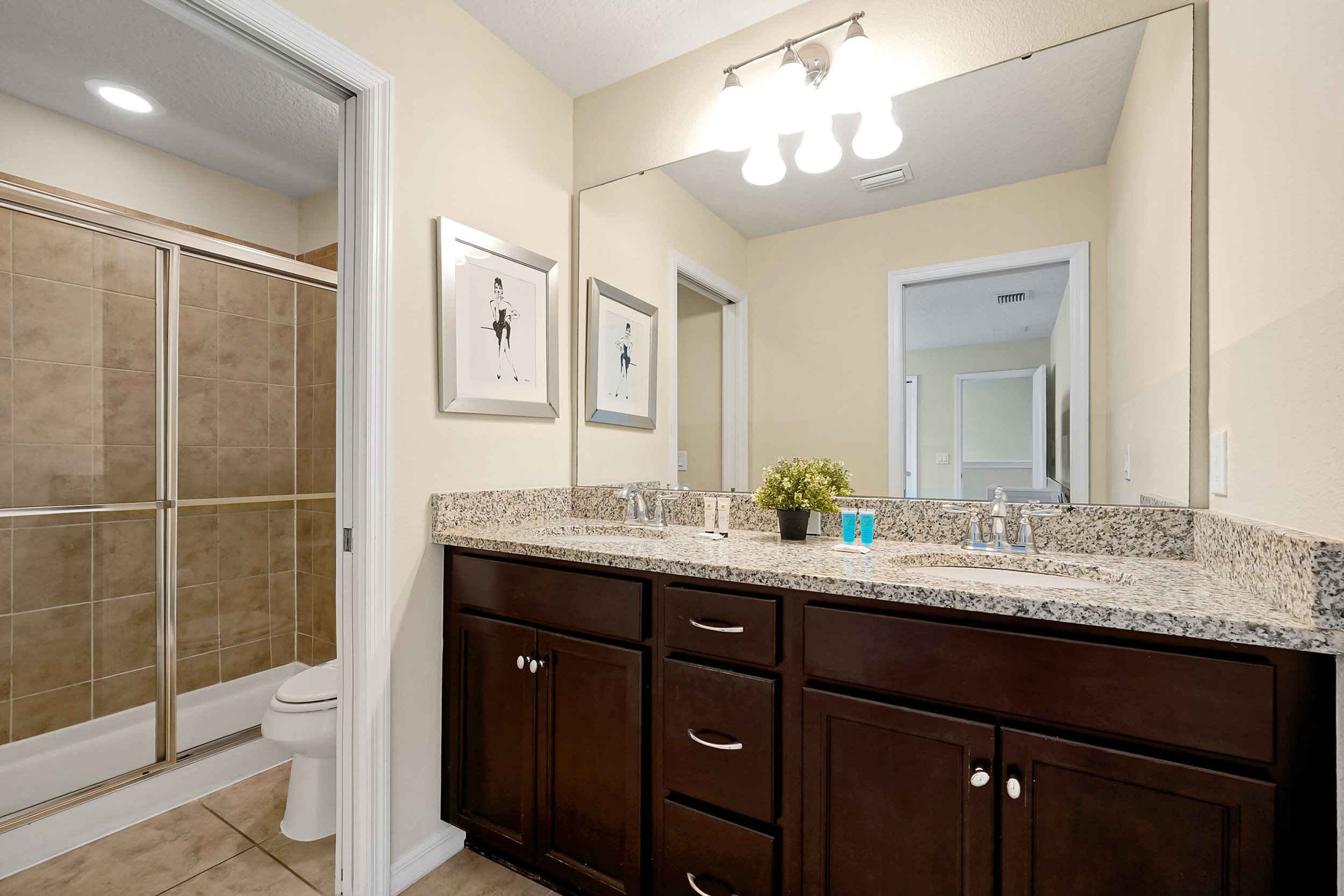 Bathroom with dark wood vanity, granite countertop, large mirror, wall-mounted light fixture, framed artwork of a woman, tiled shower, and beige walls.