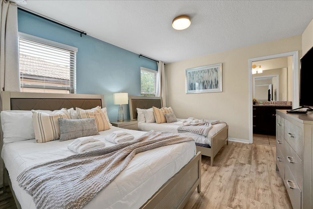 A hotel room with two queen beds, light-colored furniture, a dresser with a television, a bedside lamp, and window blinds, featuring light-colored walls with one blue accent wall.