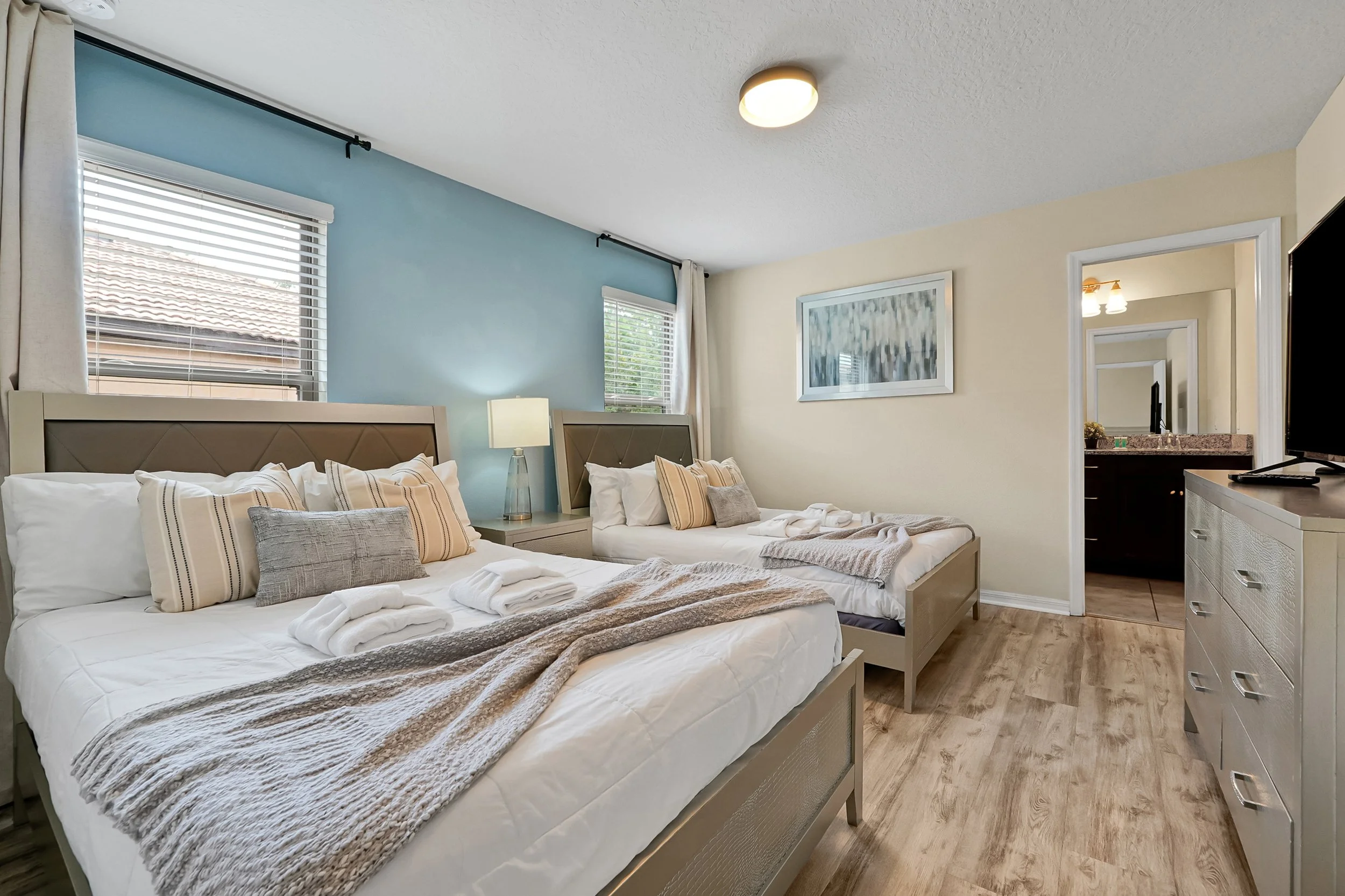 A hotel room with two beds, each with white and beige striped bedding and pillows, and a small nightstand with a lamp between them. There are two large windows with white blinds, and a piece of abstract wall art above the beds. The room has light-col