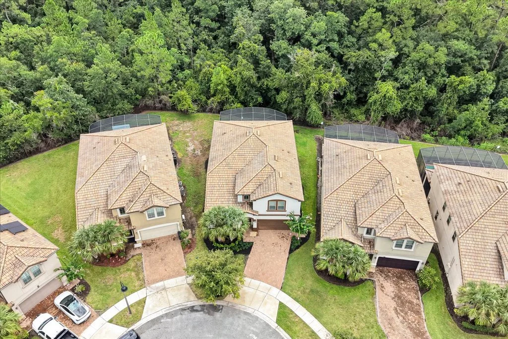 Aerial view of three suburban houses with tan roofs, green lawns, and palm trees, surrounded by lush forest; each house has a screened porch and a driveway.