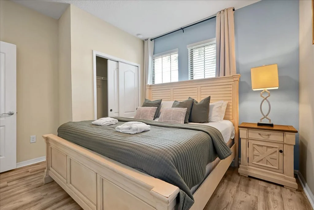 A neatly made bedroom with a wooden bed frame, green bedding, multiple pillows, a nightstand with a lamp, and a closet with sliding doors.