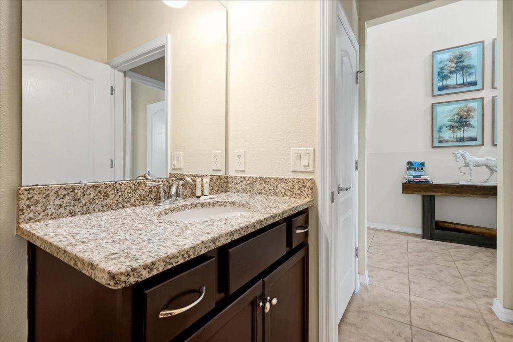 Bathroom vanity with granite countertop and sink, mirror, and wall outlets; view into adjacent room with decorative horse sculpture and framed pictures of trees on the wall.