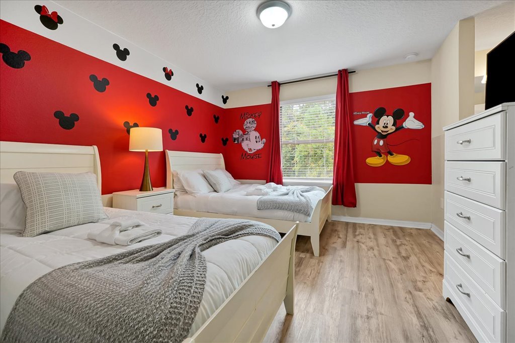 Bedroom with red Mickey Mouse themed wallpaper, featuring black Mickey head silhouettes and a large painted image of Mickey Mouse on the wall, with white beds, a nightstand, a lamp, and a window with red curtains.