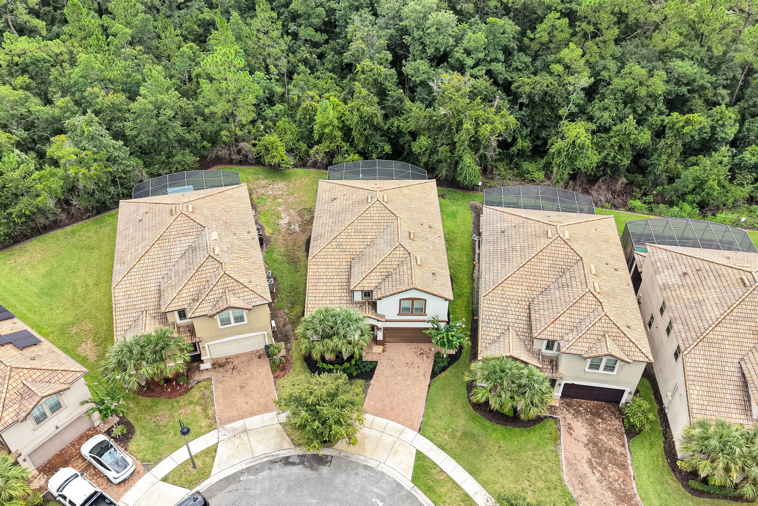 An aerial view of several suburban houses with tile roofs, surrounded by green lawns, trees, and a forested area in the background. A curved street and parked cars are visible in the foreground, along with screened-in porches on some of the houses.