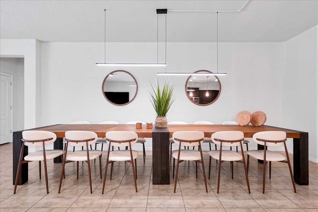 Modern dining room with a long wooden table, white chairs, a green plant centerpiece, two round mirrors, and decorative spheres on the table. The room has white walls, tiled flooring, and minimalist lighting fixtures.