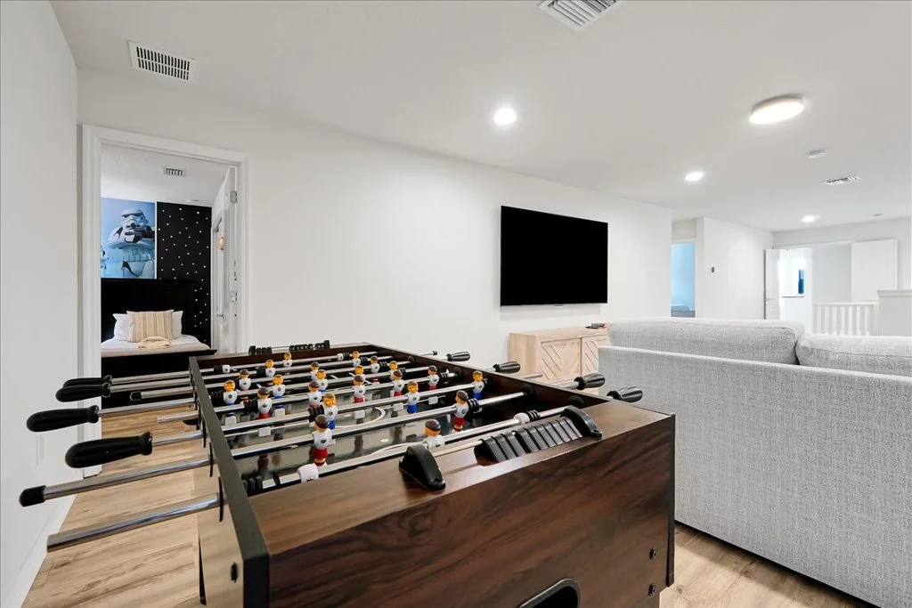 A spacious living room with a foosball table, a gray sofa, a wall-mounted flat-screen TV, and a small bedroom visible through an open door in the background.