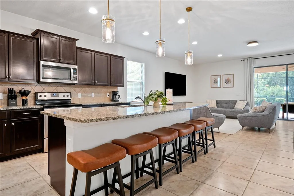 Open-concept kitchen and living room with dark wood cabinets, granite countertops, white walls, and tiled floor. Kitchen features microwave, stove, coffee maker, and barstools. Living area has gray furniture, wall-mounted TV, framed artwork, and slid
