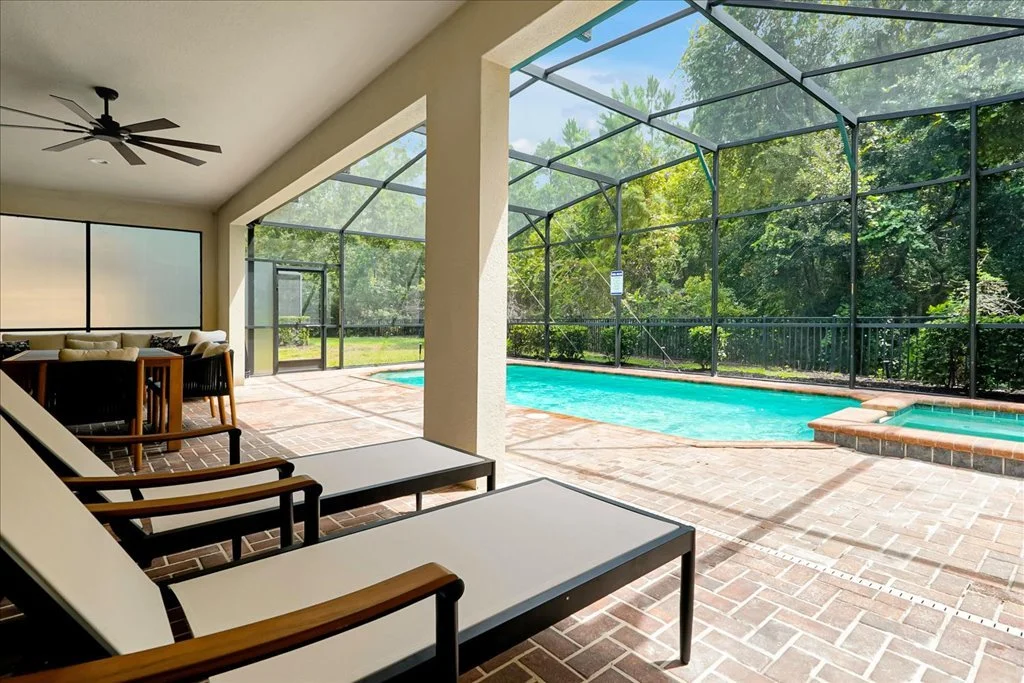Covered patio area with lounge chairs, a pool, and a screened enclosure with trees outside.