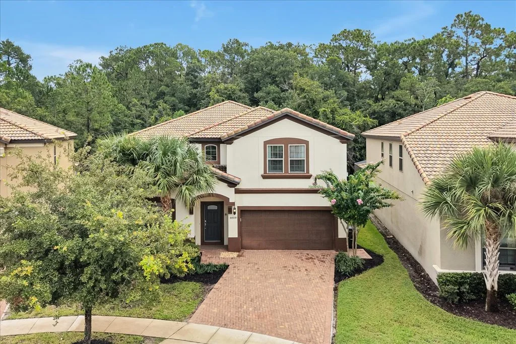 Two-story house surrounded by trees and bushes, with a paved driveway and a landscaped front yard.