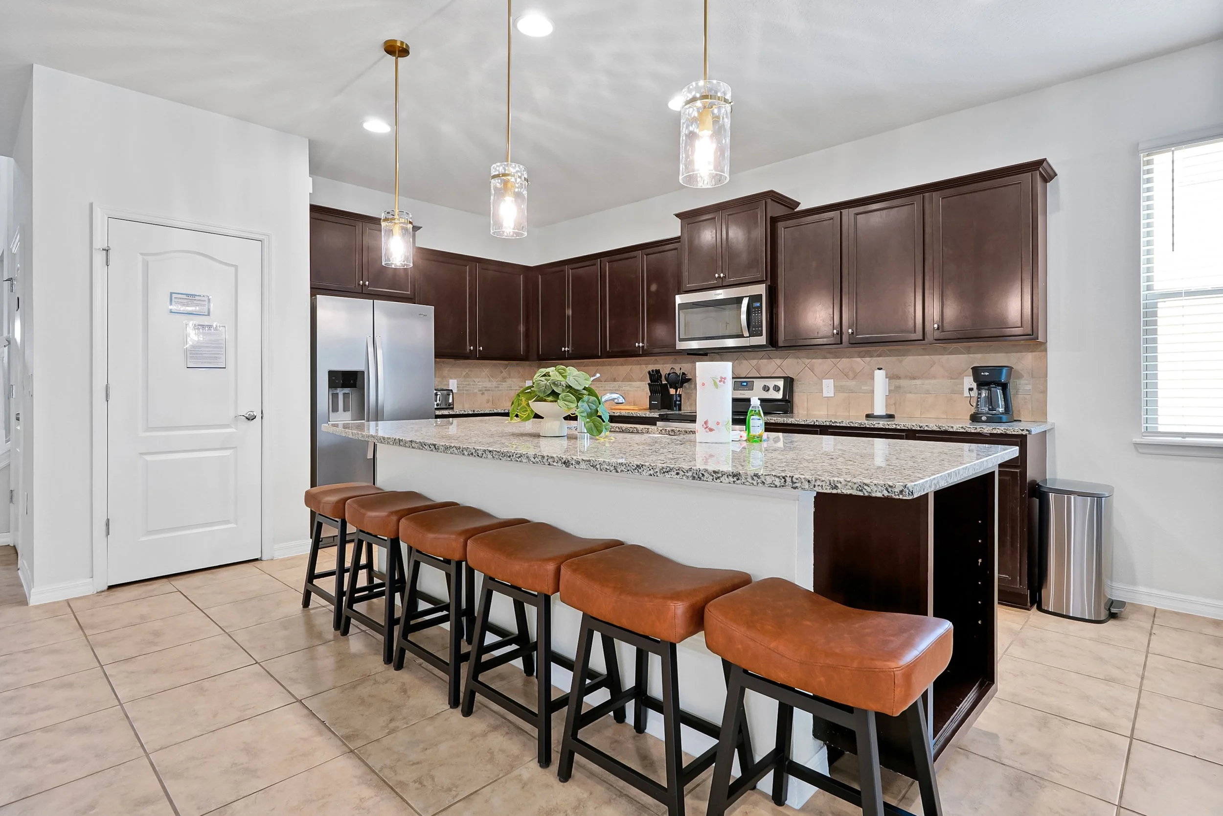 Modern kitchen with dark wood cabinets, stainless steel microwave, and refrigerator, cream-colored granite counter, white walls, pendant lights, and bar stools along the kitchen island, beige tile floor.