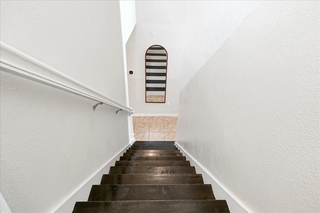 View of a staircase with dark wooden steps and a white wall on the right, leading down to a small landing with beige tile, a mirror with a brown frame on the wall at the bottom, and a staircase visible in the mirror.