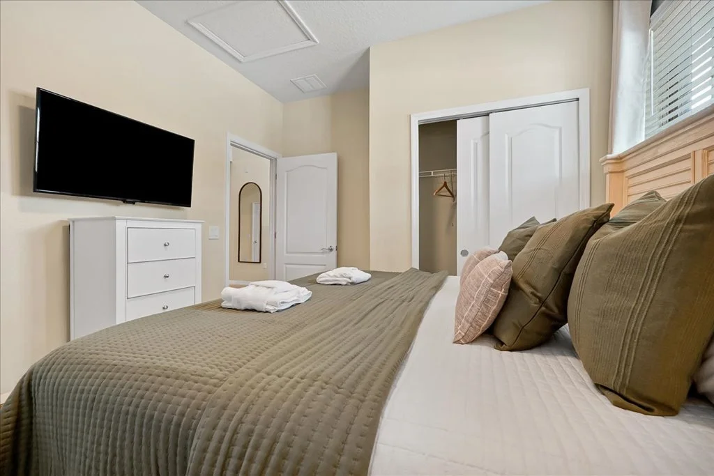 A bedroom with a bed, multiple pillows, a wall-mounted TV, a dresser, a closet with open doors, and a window with blinds.