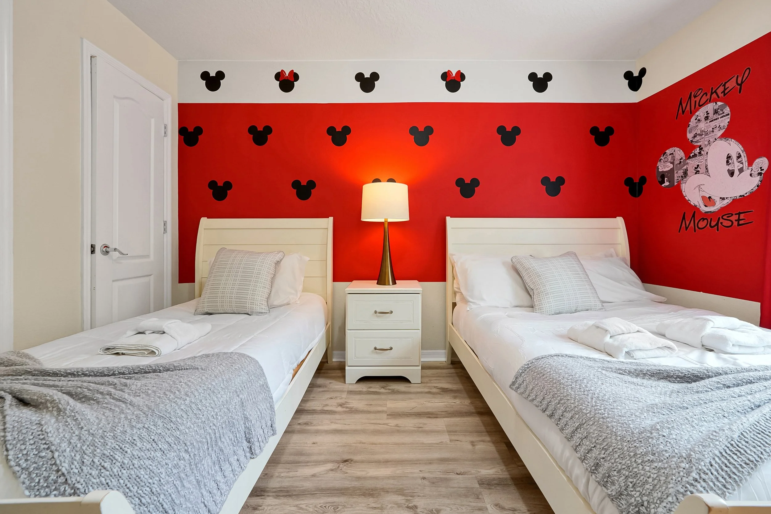 A bedroom with two twin beds, white headboards, gray blankets, and pillows, a white nightstand with a lamp, and a wall decorated with Mickey Mouse silhouettes and a Mickey Mouse graphic with the text "Mickey Mouse" on a red and white background.