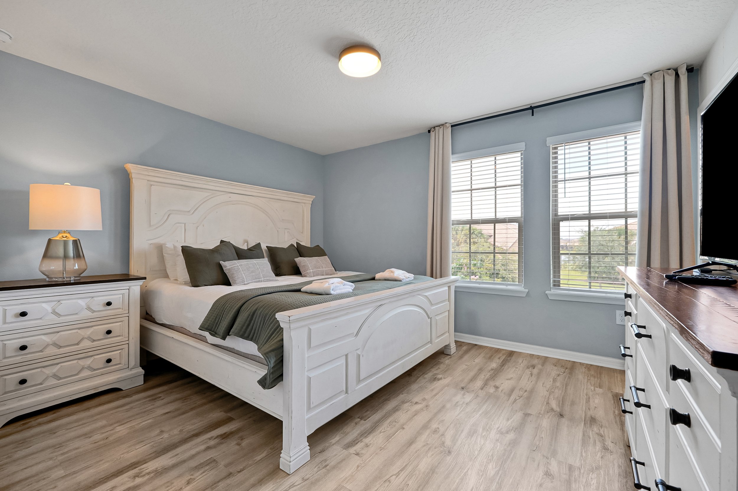 A bedroom with light blue walls, a white wooden bed with a headboard, dark green and light gray pillows, a nightstand with a lamp, and large windows with white blinds and beige curtains, overlooking trees and rooftops outside.