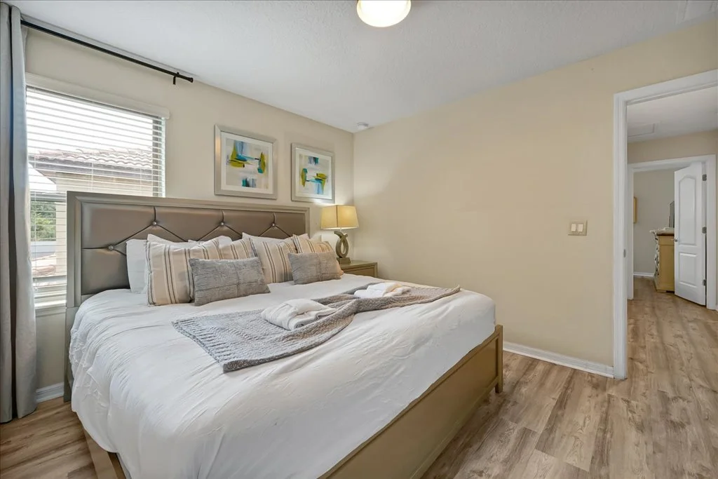 A neatly made bed with striped pillows and a folded blanket, a bedside table with a lamp, and abstract artwork above the bed in a cozy bedroom with beige walls and wood flooring.