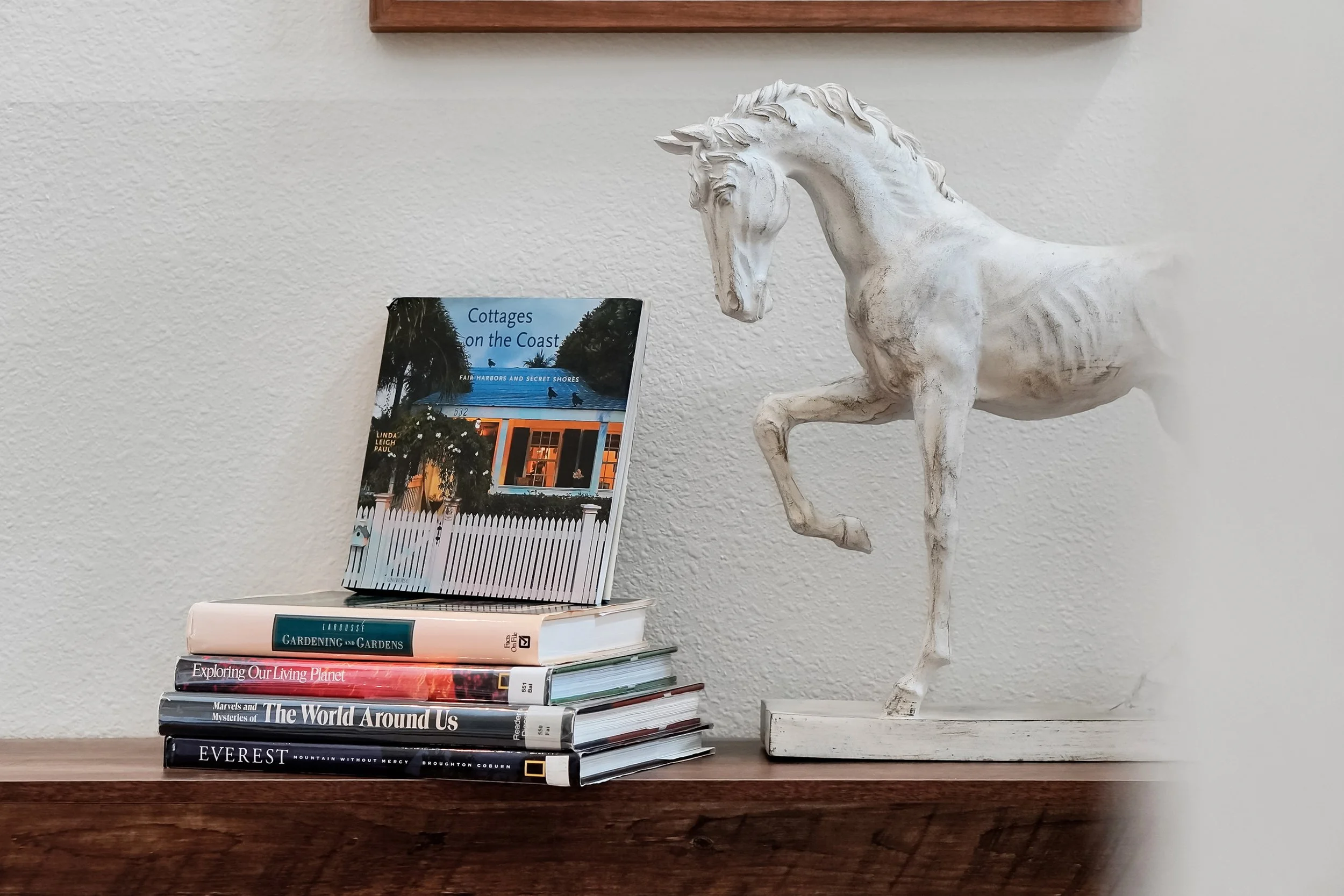 A white ceramic horse sculpture on a wooden table next to a stack of books and a magazine on a white wall background.
