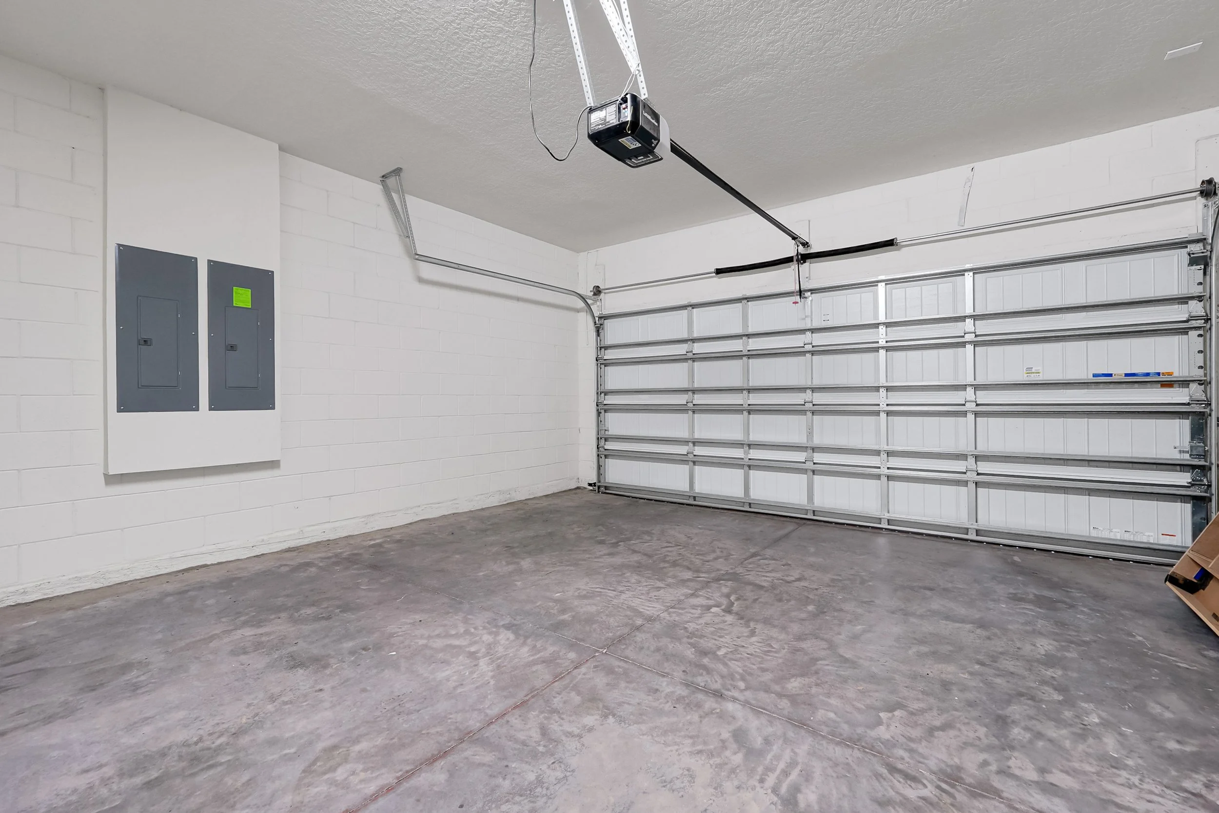 Empty garage with a closed metal roll-up door, concrete floor, white brick walls, and an electric panel on the wall.