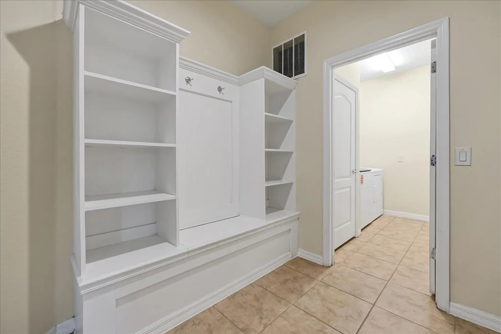 Empty white built-in bookshelf and cabinet unit against beige wall in a room with tiled floor, doorway leading to laundry area with washing machine, and a vent on the wall.