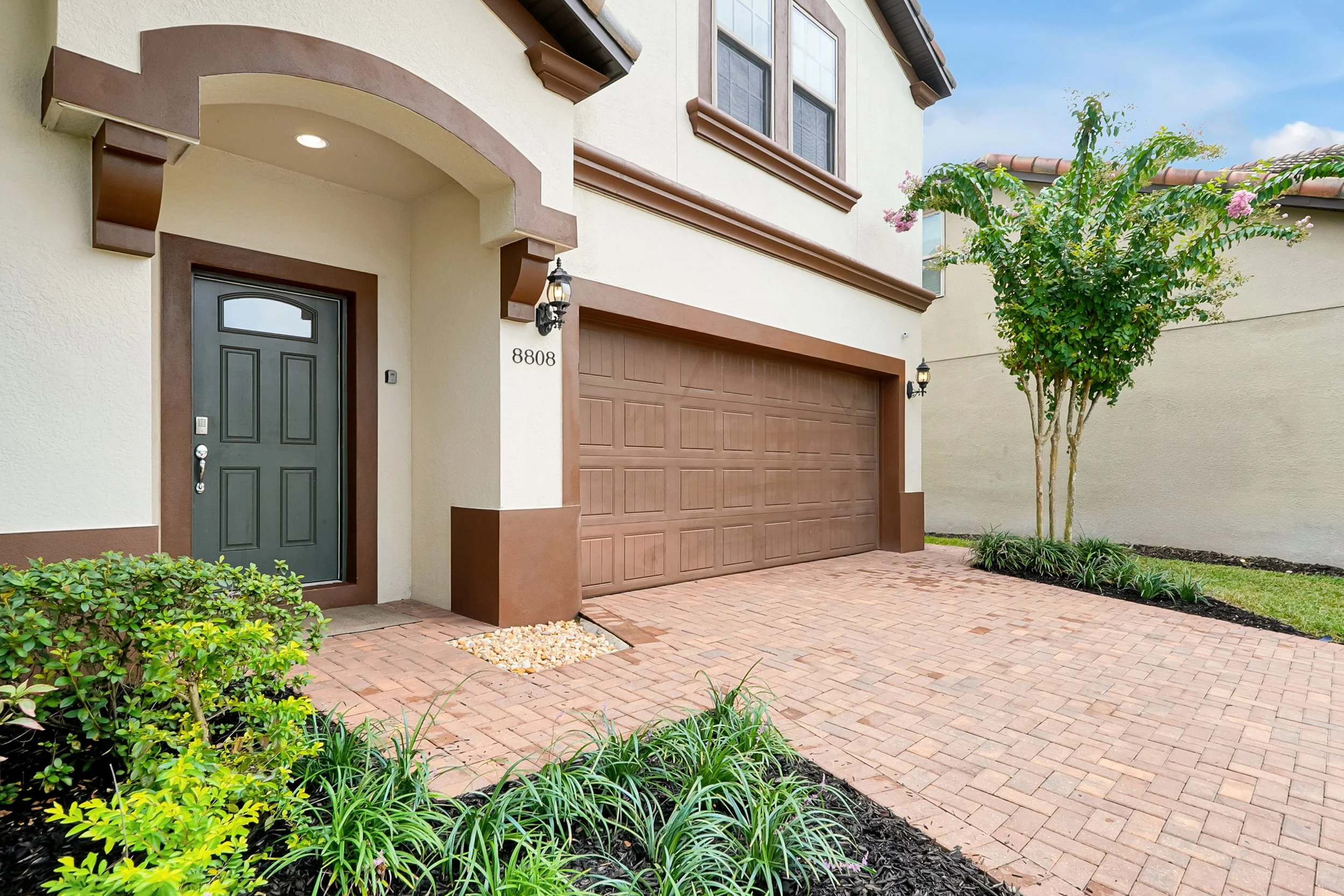 Front of a two-story house with beige walls and brown trim, a green front door, a brown garage door, and a small landscaped yard with bushes and a tree.