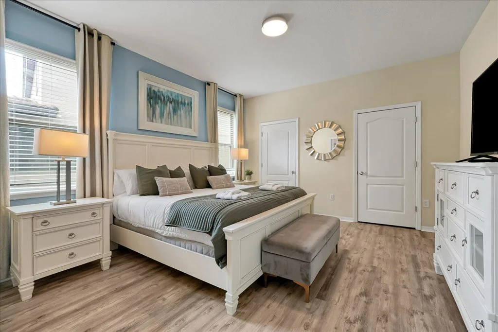 A bedroom with a white bed, multiple dark and light pillows, beige and blue walls, two nightstands with lamps, a painting above the bed, a gray bench at the foot of the bed, a white dresser with a TV, a round mirror, and two white doors.
